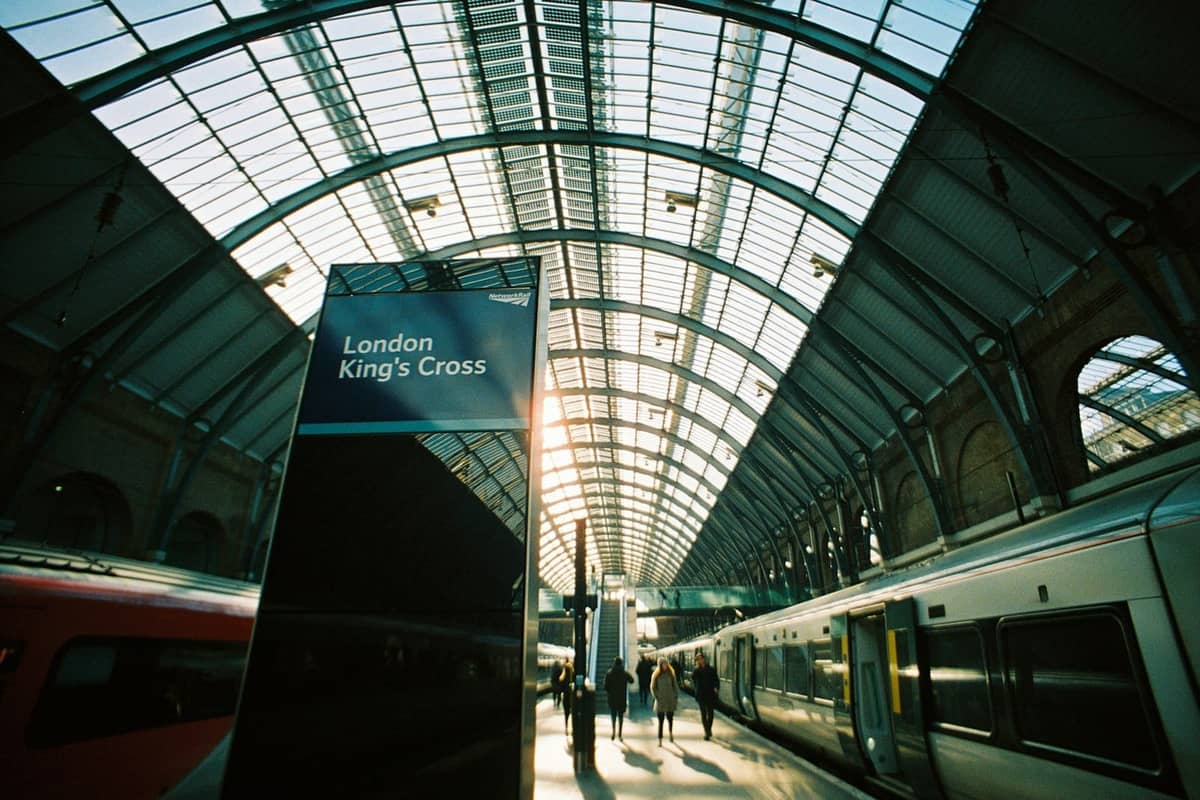 Interior da estação ferroviária London King's Cross com telhado arqueado de vidro, trens nas plataformas e uma placa com os dizeres 'London King's Cross'.