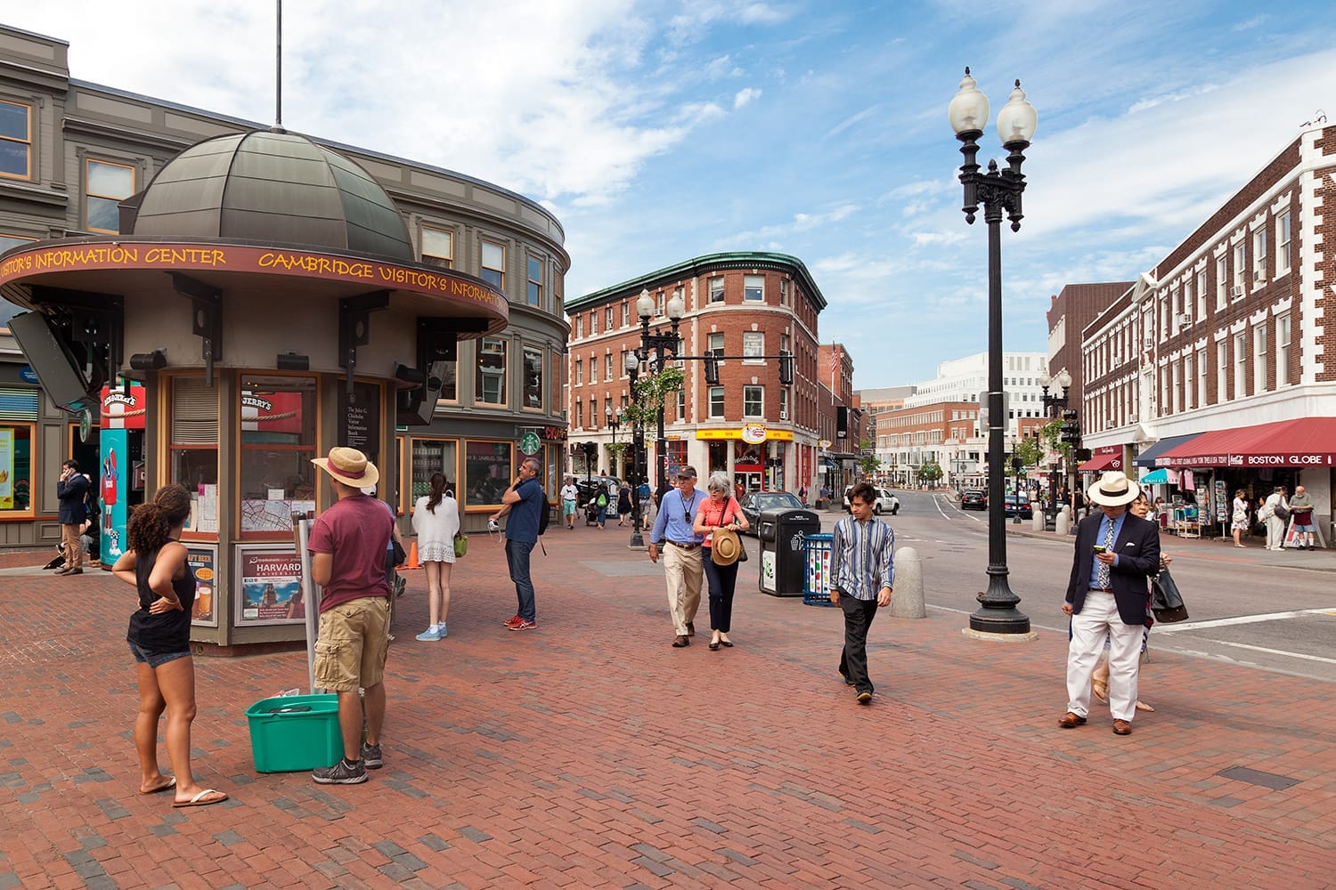 Persone camminano e si riuniscono vicino al Cambridge Visitor's Information Center a Harvard Square, circondate da storici edifici in mattoni sotto un cielo parzialmente nuvoloso.