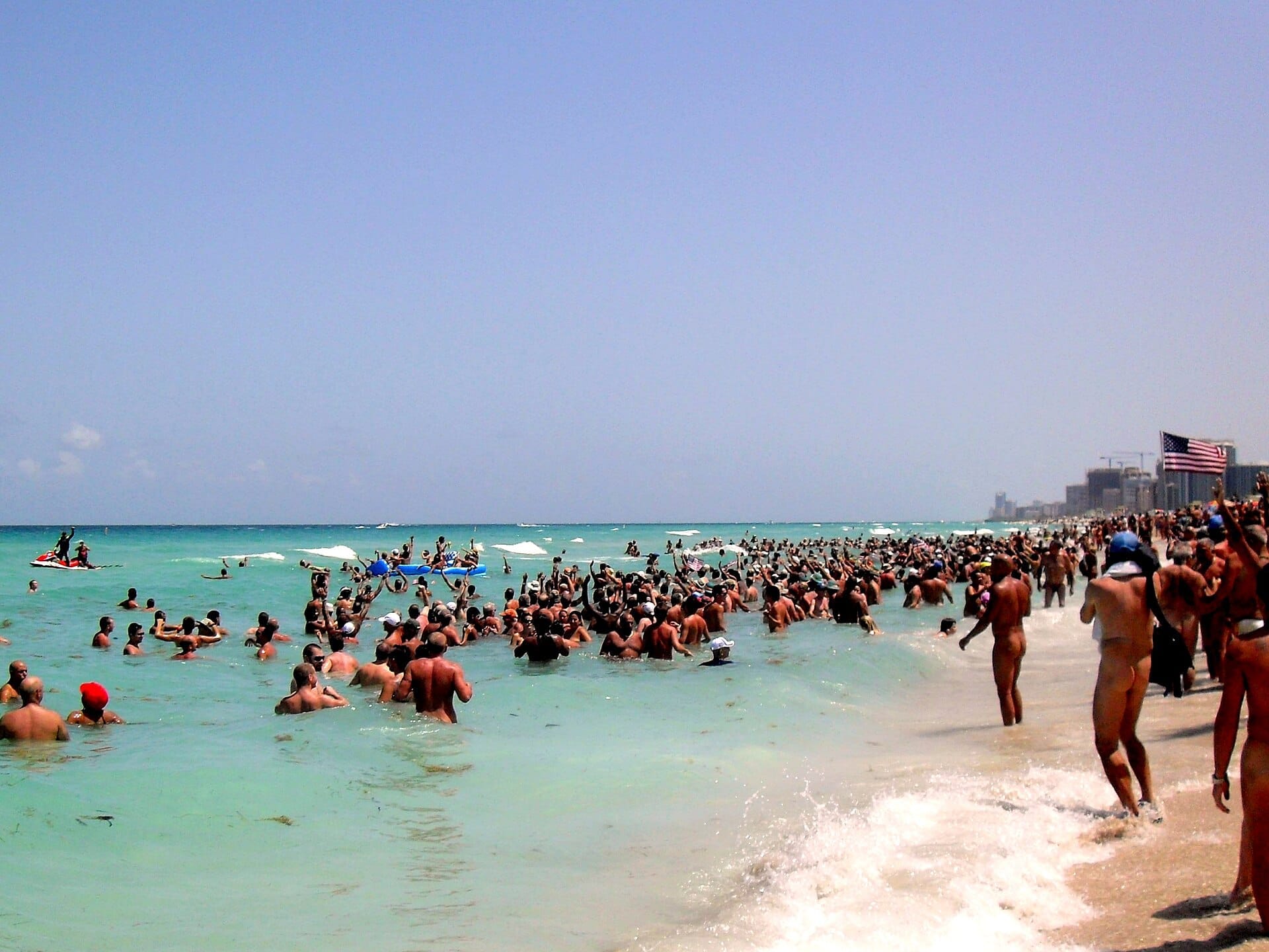 Le folle godono dell'acqua turchese limpida e della riva sabbiosa a Haulover Beach Park sotto un cielo azzurro brillante, con gli edifici della città in lontananza.