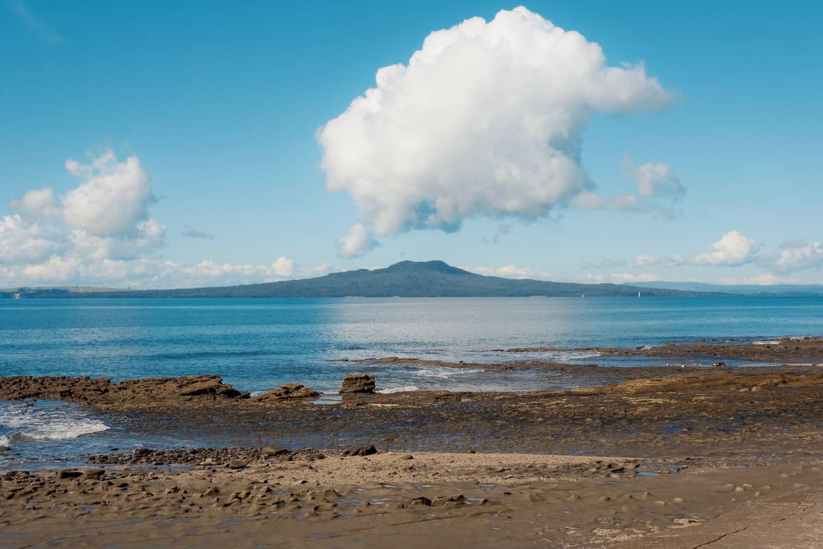 Widok przez wodę na wyspę Rangitoto, charakterystyczny stożek wulkaniczny pod błękitnym niebem i białymi chmurami, widziany z brzegu Auckland.