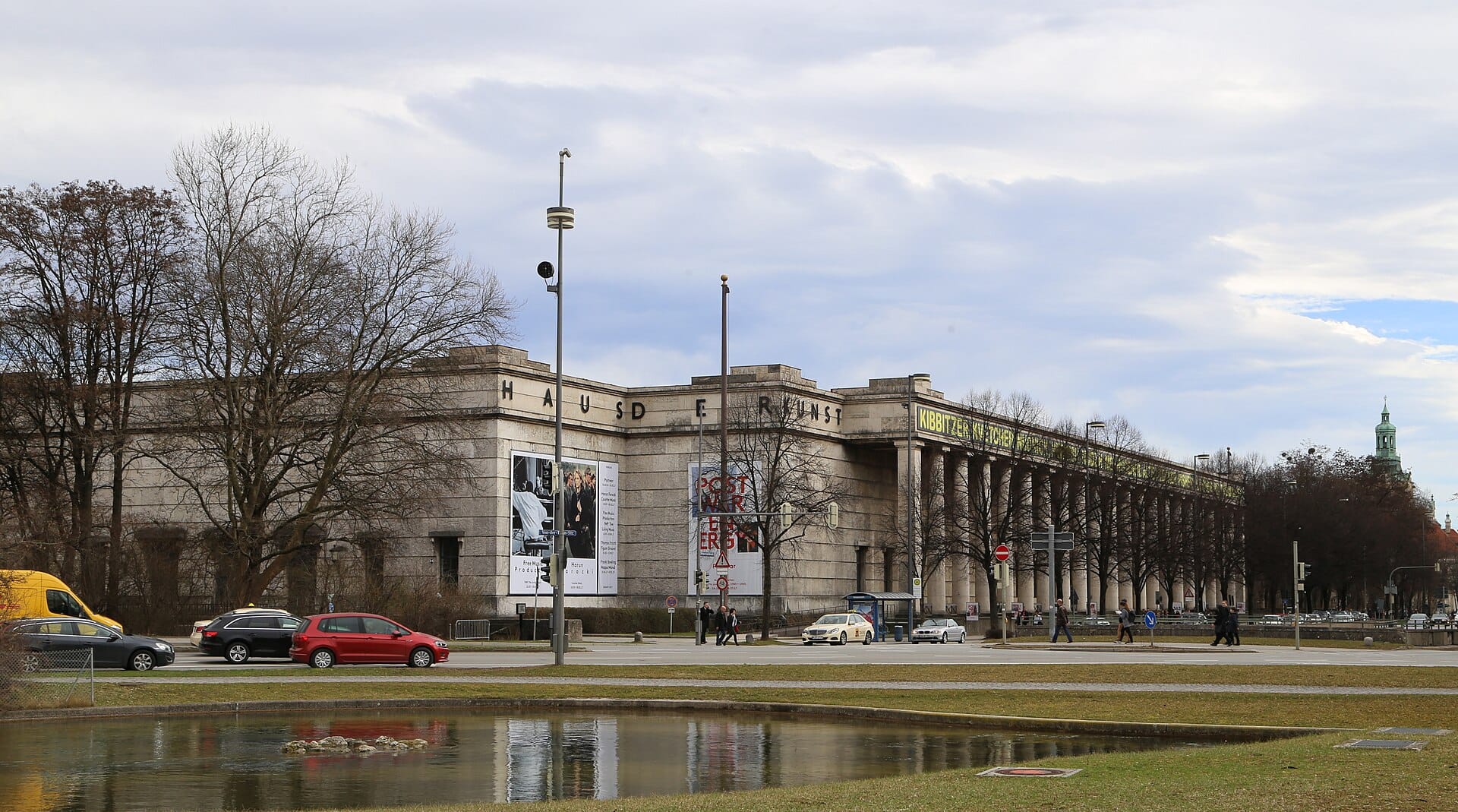 ภาพมุมกว้างของหน้าตึก Haus der Kunst มิวนิก อาคารนีโอคลาสสิกขนาดใหญ่ เสาสูง โปสเตอร์สีสด มองจากข้ามถนน
