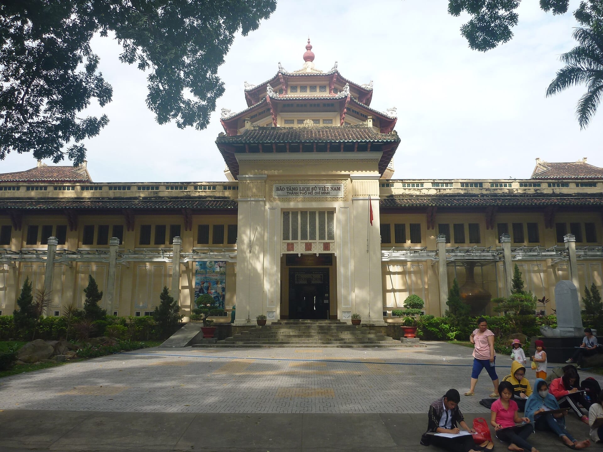 Vista frontal del Museo de Historia de Ho Chi Minh con su fachada colonial, tejados de pagoda y visitantes sentados en el patio de entrada sombreado.