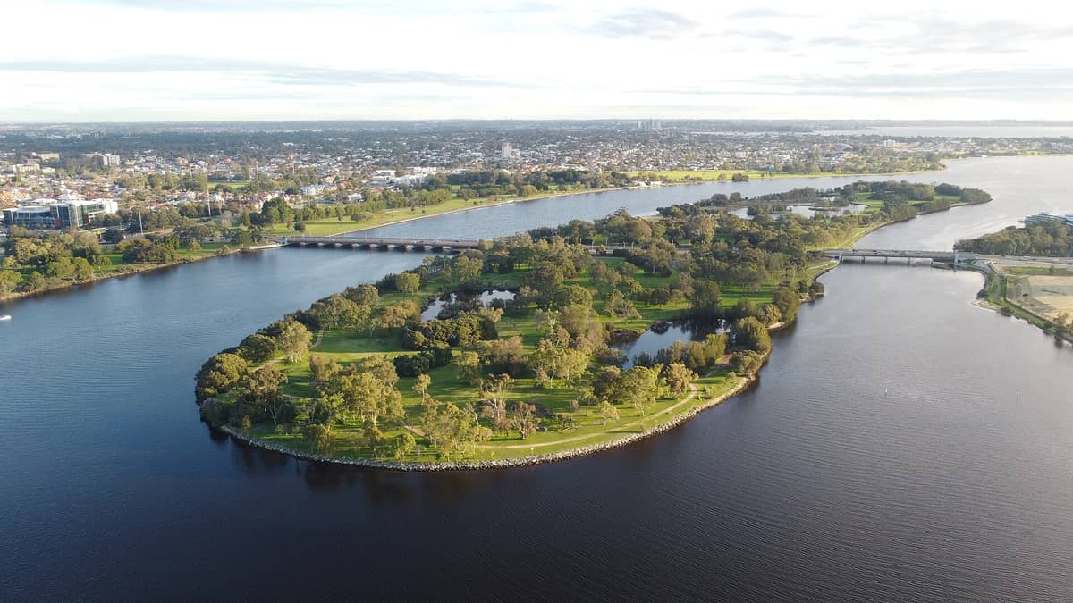Luftaufnahme von Heirisson Island mit dem Swan River, umliegenden Parkanlagen, Brücken und der Skyline von Perth im Hintergrund unter einem teils bewölkten Himmel.