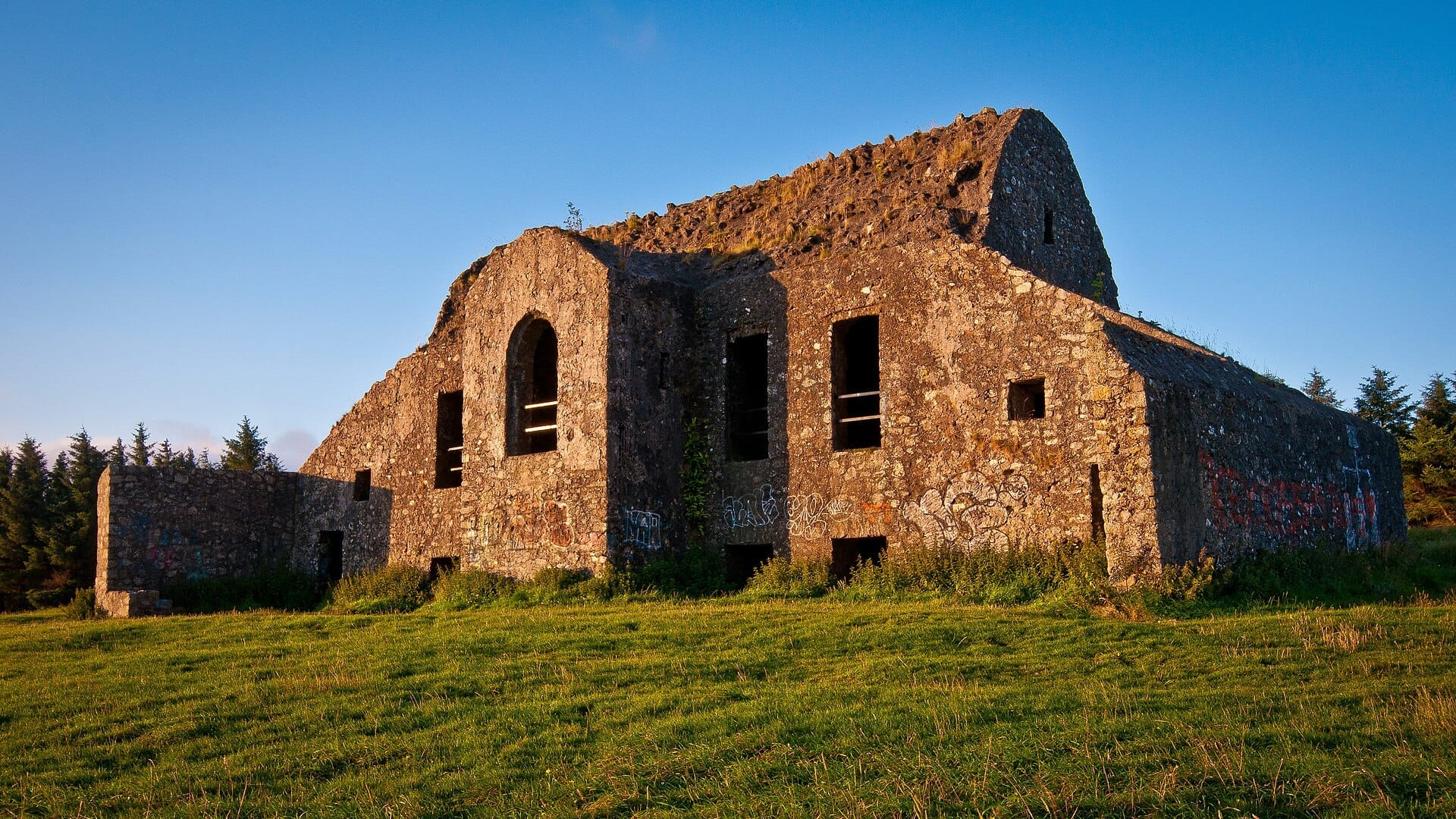 Il rudere dell'Hellfire Club si trova sul verde Montpelier Hill sotto un cielo azzurro limpido, circondato da alberi sempreverdi lontani e dalla luce dorata della sera.