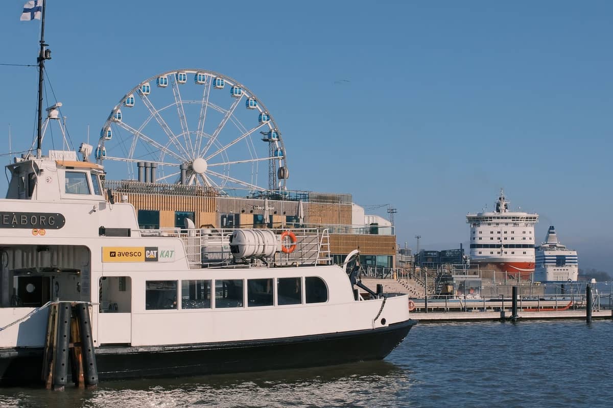 Cena à beira-mar em Helsinki com balsa, roda-gigante e paisagem urbana incluindo navios e edifícios portuários.