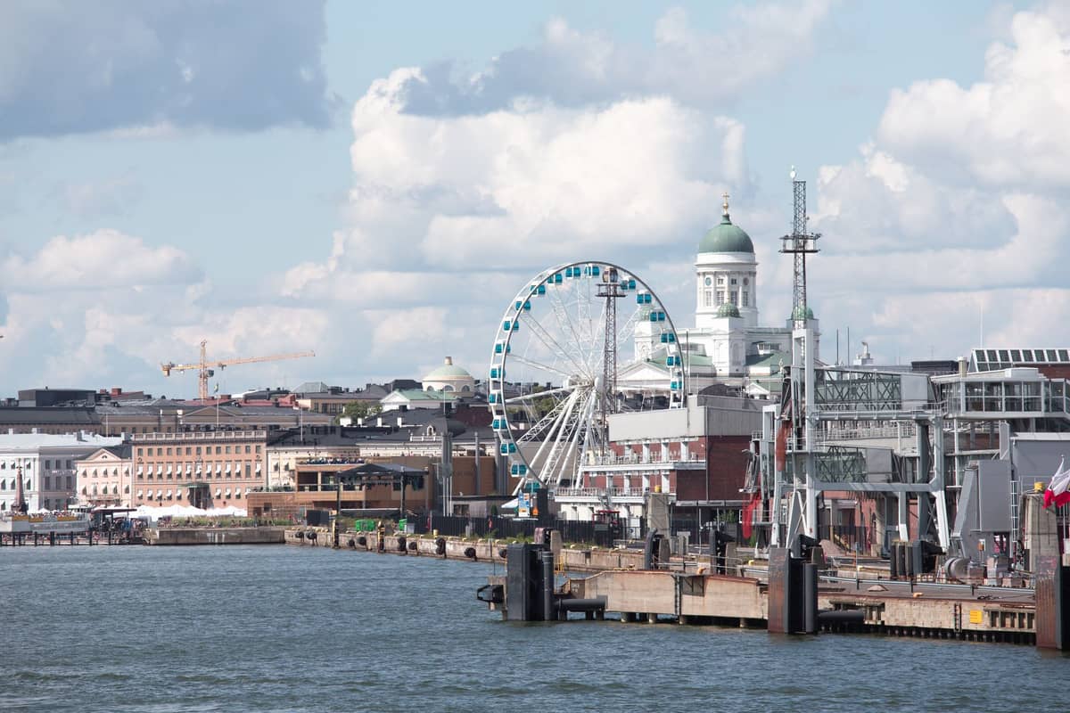Vista ampla da orla de Helsinque com a SkyWheel, a cúpula da Catedral de Helsinque e as estruturas históricas do porto sob um céu azul com nuvens.