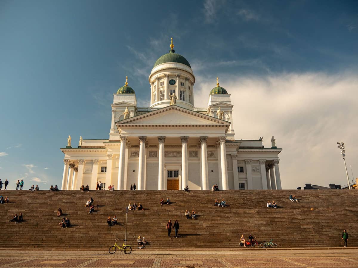 Vista ampla da Catedral de Helsinque com suas altas cúpulas verdes erguendo-se acima de uma grande escadaria, pessoas reunidas nos degraus sob um céu azul.