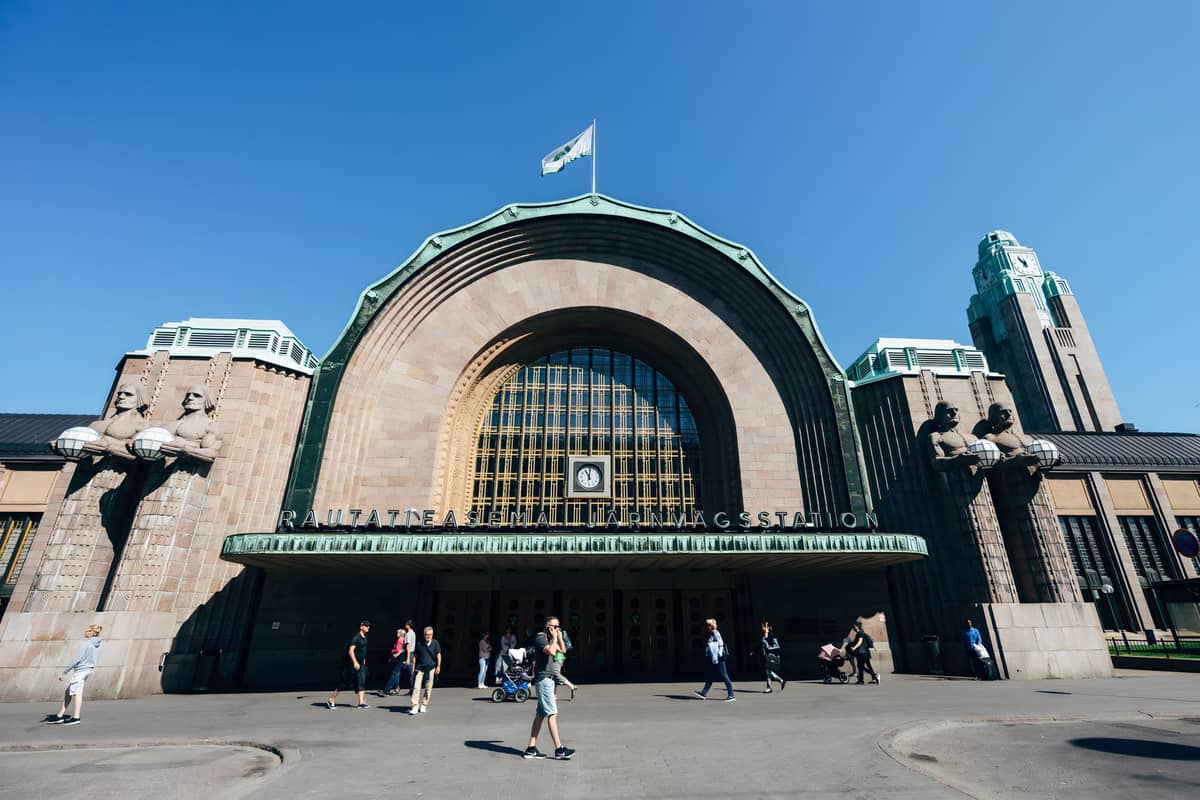 Vista frontal ampla da Estação Central de Helsinque mostrando sua icônica entrada em arco, as figuras de pedra segurando lanternas, a torre do relógio e pessoas do lado de fora em um dia ensolarado.