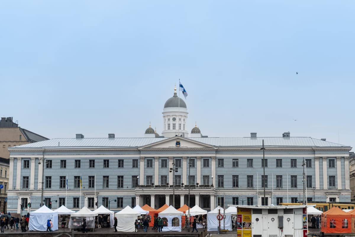 Barracas do mercado de Natal montadas em frente à Câmara Municipal de Helsinque, perto da Praça do Senado, com a cúpula do edifício e a bandeira finlandesa visíveis no inverno.