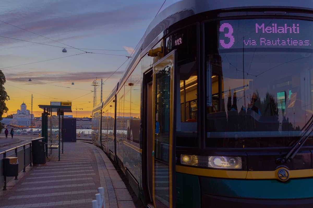 Eléctrico moderno de Helsínquia numa paragem da cidade com a Catedral de Helsínquia visível ao fundo ao pôr do sol, mostrando o acesso ao transporte público.