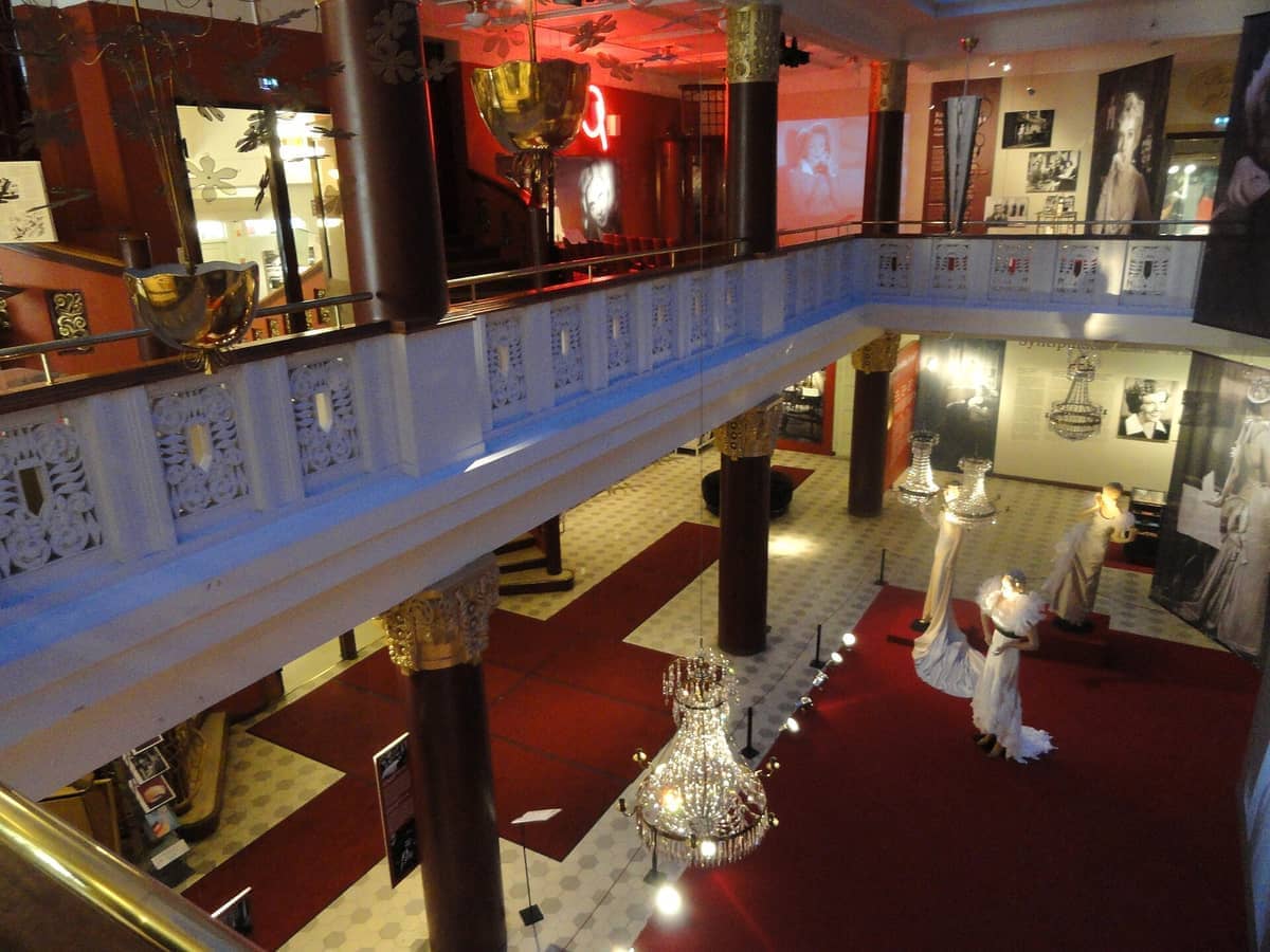 Vista interior do Helsinki City Museum com lustres ornamentados, manequins em trajes de época, carpete vermelho e fotografias históricas nas paredes.