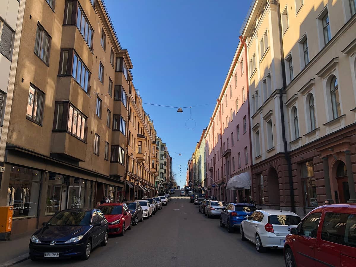 Vista de uma rua no Helsinki Design District mostrando edifícios históricos coloridos, carros estacionados e um céu azul claro numa tarde ensolarada.