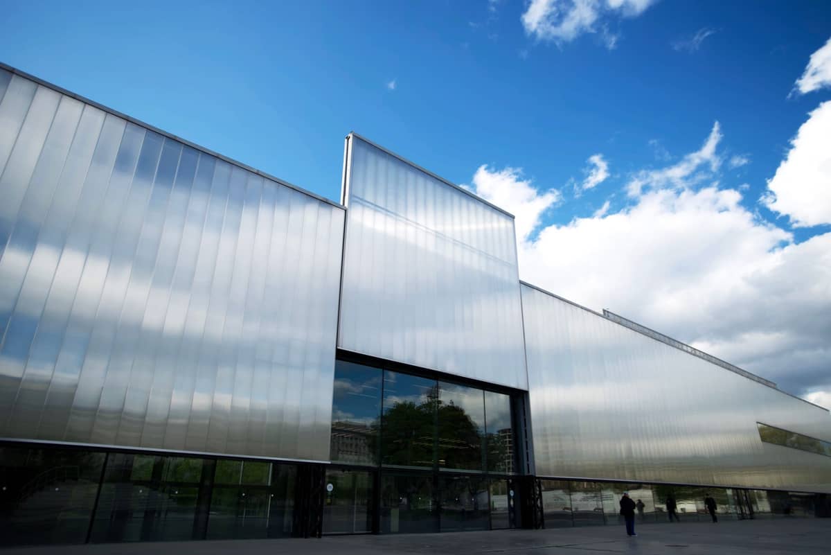 Moderno edifício de museu em vidro e metal com fachada geométrica sob céu azul, ampla calçada à frente e reflexo de árvores próximas em Helsinki.