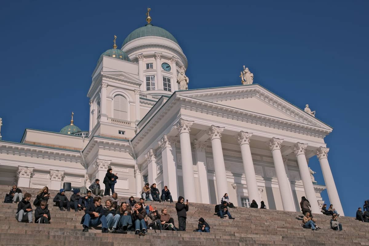 Pessoas sentadas e caminhando nos amplos degraus de pedra em frente à Catedral de Helsinque, uma igreja neoclássica branca sob um céu azul.