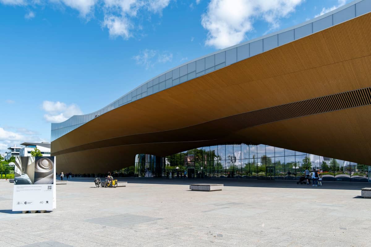 Vista ampla da Biblioteca Central Oodi de Helsinque, um marcante edifício moderno com telhado curvo de madeira e fachada de vidro, em um dia ensolarado.