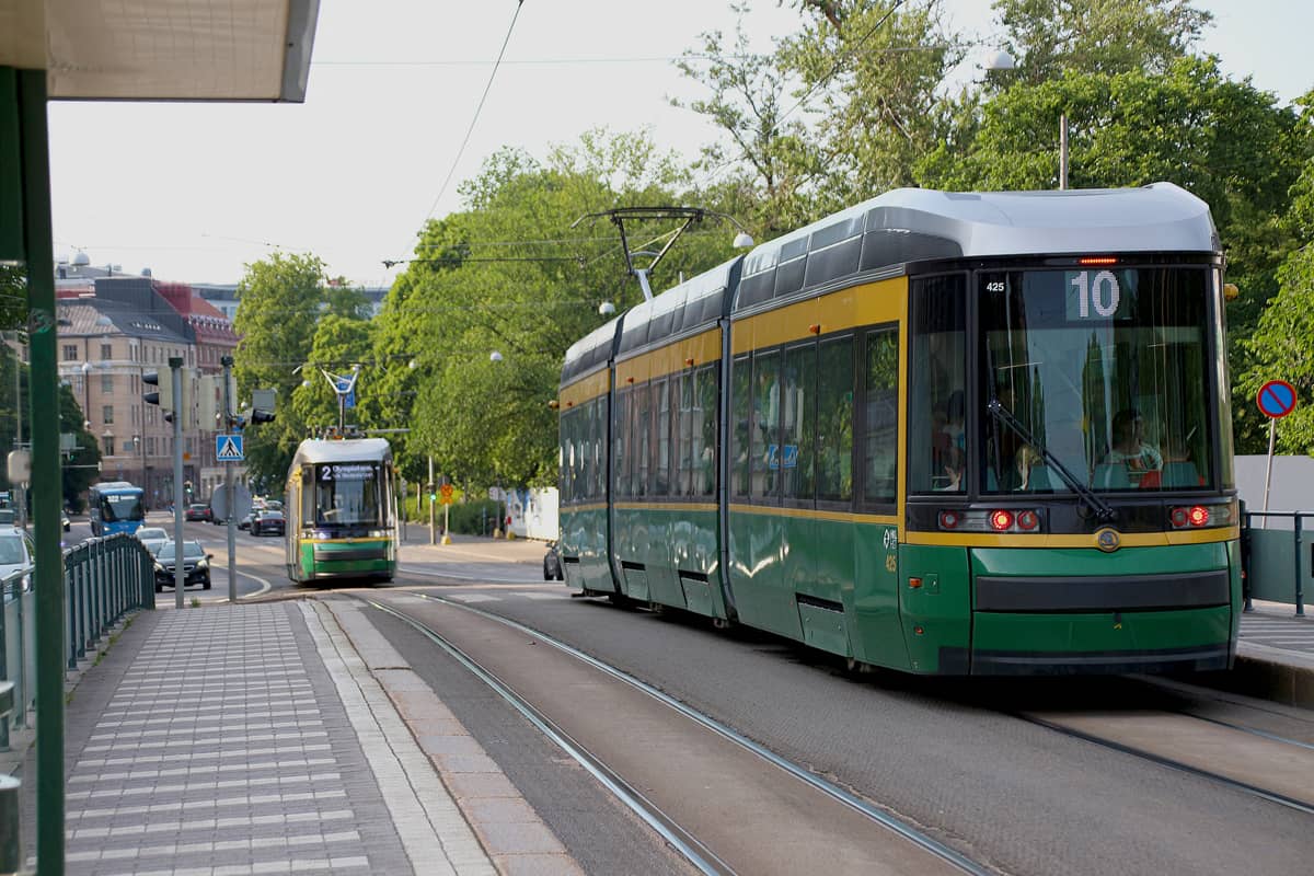Modernos elétricos verdes de Helsinque passando por uma rua arborizada e uma paragem de elétrico num luminoso dia de verão