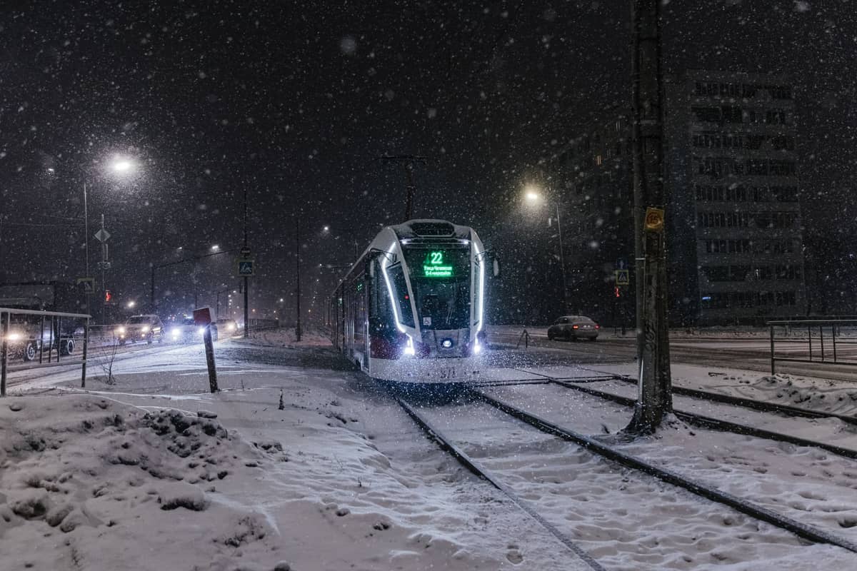Bonde moderno percorrendo as ruas nevadas de Helsinki à noite, iluminado por postes de luz e cercado por neve fresca nos trilhos e calçadas.