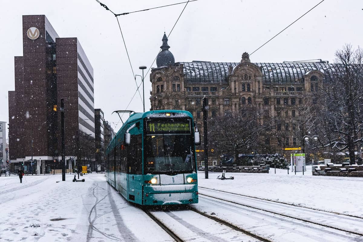 Um bonde turquesa percorre trilhos cobertos de neve no centro de Helsinque, cercado por edifícios históricos e modernos durante uma nevada diurna.