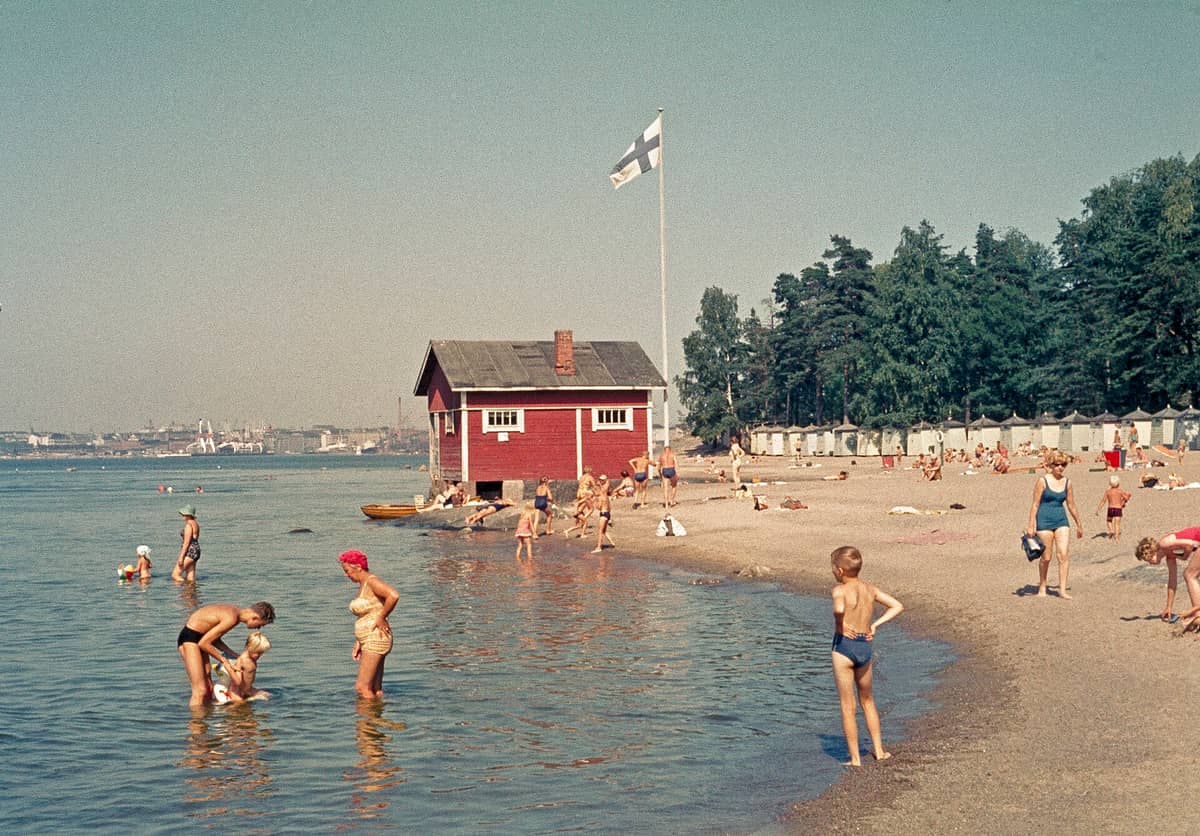 Ilha de praia de areia em Helsinki com pessoas nadando, uma sauna de madeira vermelha, bandeira finlandesa, pinheiros e cabanas de praia coloridas sob o sol de verão.