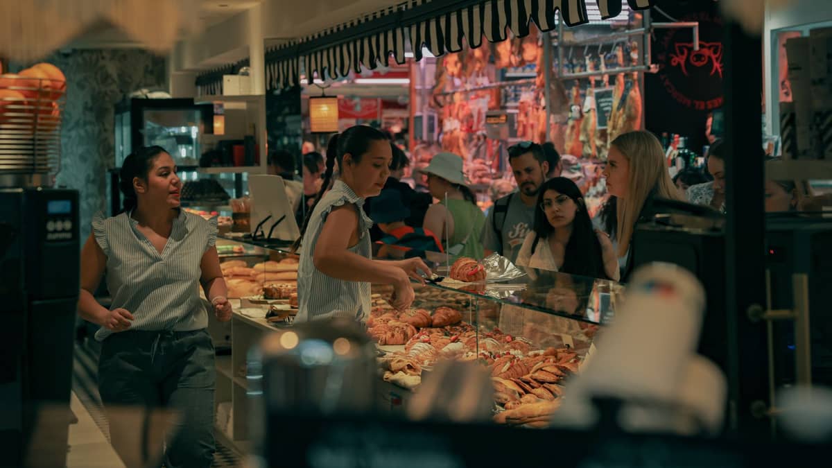Um movimentado balcão de mercado coberto com funcionários atendendo clientes e uma exposição de produtos de padaria, doces e pães em um mercado interno de estilo nórdico.