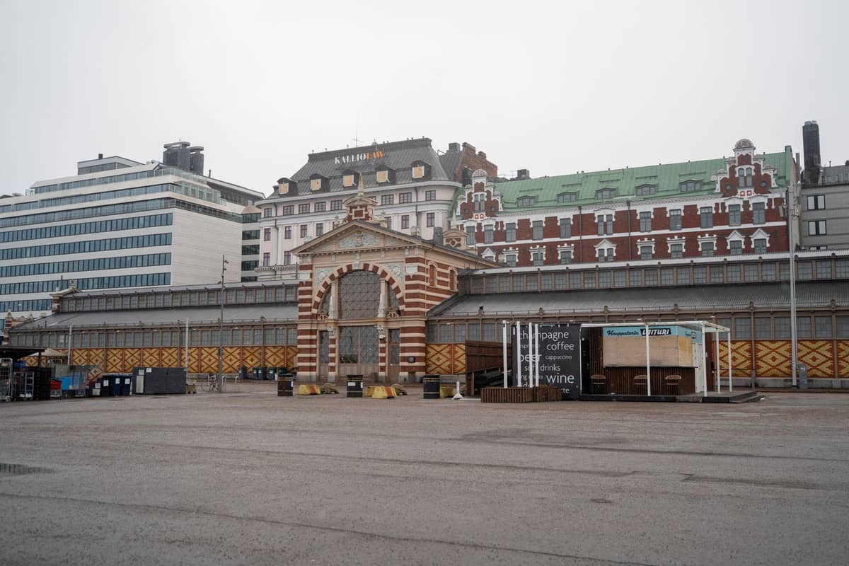 O exterior de um histórico edifício de mercado coberto em Helsinki com alvenaria ornamentada, entrada em arco e edifícios da cidade ao fundo num dia nublado.