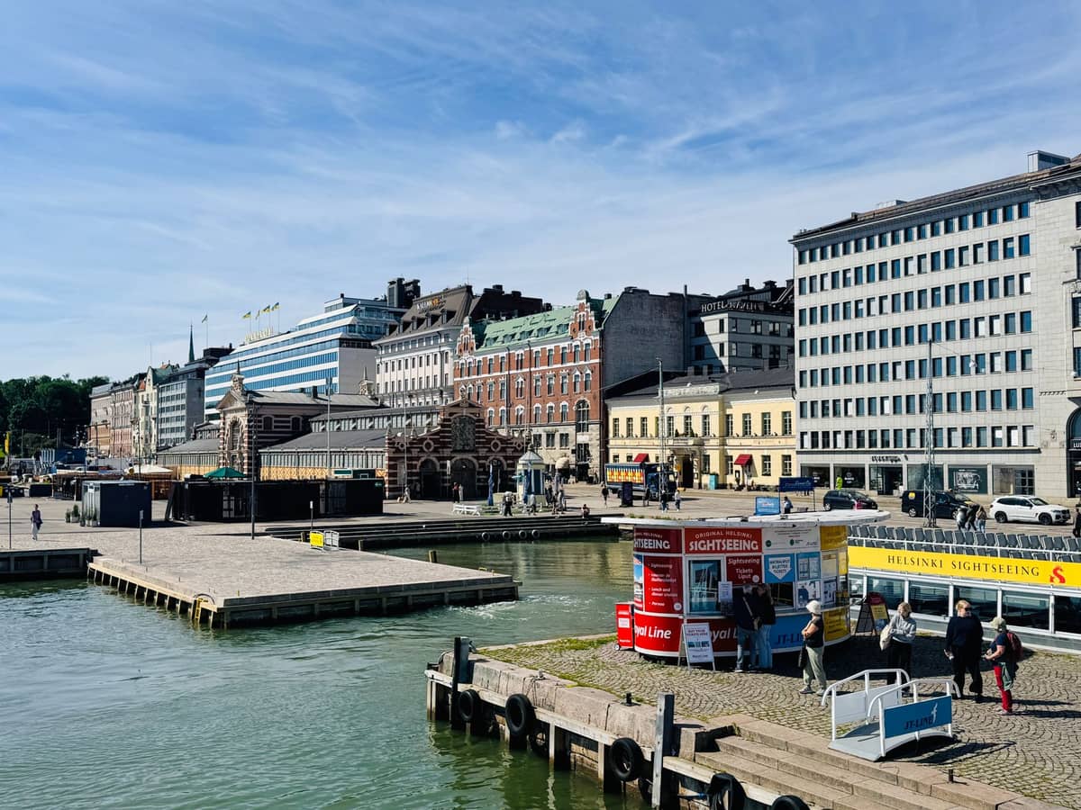 Vista do edifício do Mercado Coberto Antigo de Helsinque no porto, com os prédios históricos ao redor e a orla da cidade em um dia ensolarado.