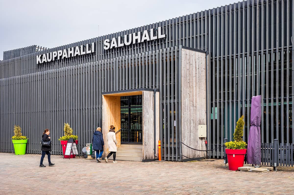 Pessoas entrando na entrada moderna do Mercado de Hakaniemi com ripas verticais distintas e grandes vasos coloridos do lado de fora.