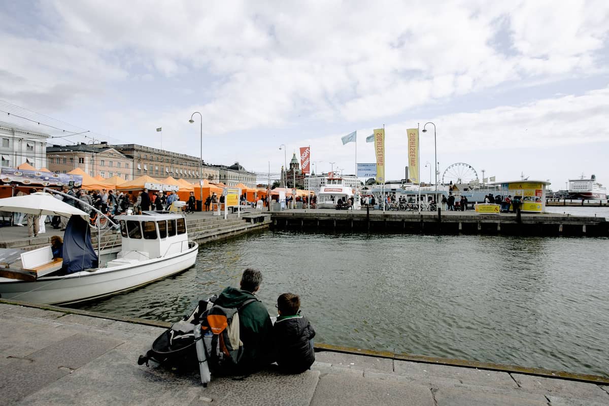 Duas pessoas sentadas à beira da água a olhar para o movimentado Porto Sul de Helsínquia, com tendas de mercado, barcos e edifícios históricos nas proximidades visíveis.