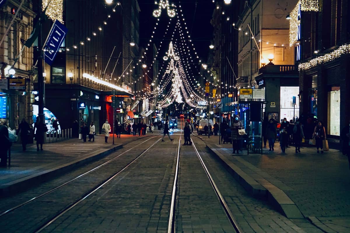 Vista noturna de uma rua movimentada no centro de Helsinque com bondes, pessoas e luzes festivas penduradas sobre a cidade.