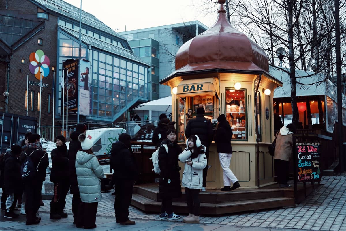 Pessoas fazem fila tranquilamente em um quiosque bar ao ar livre em uma praça da cidade, agasalhadas com roupas de inverno, com edifícios modernos ao fundo.