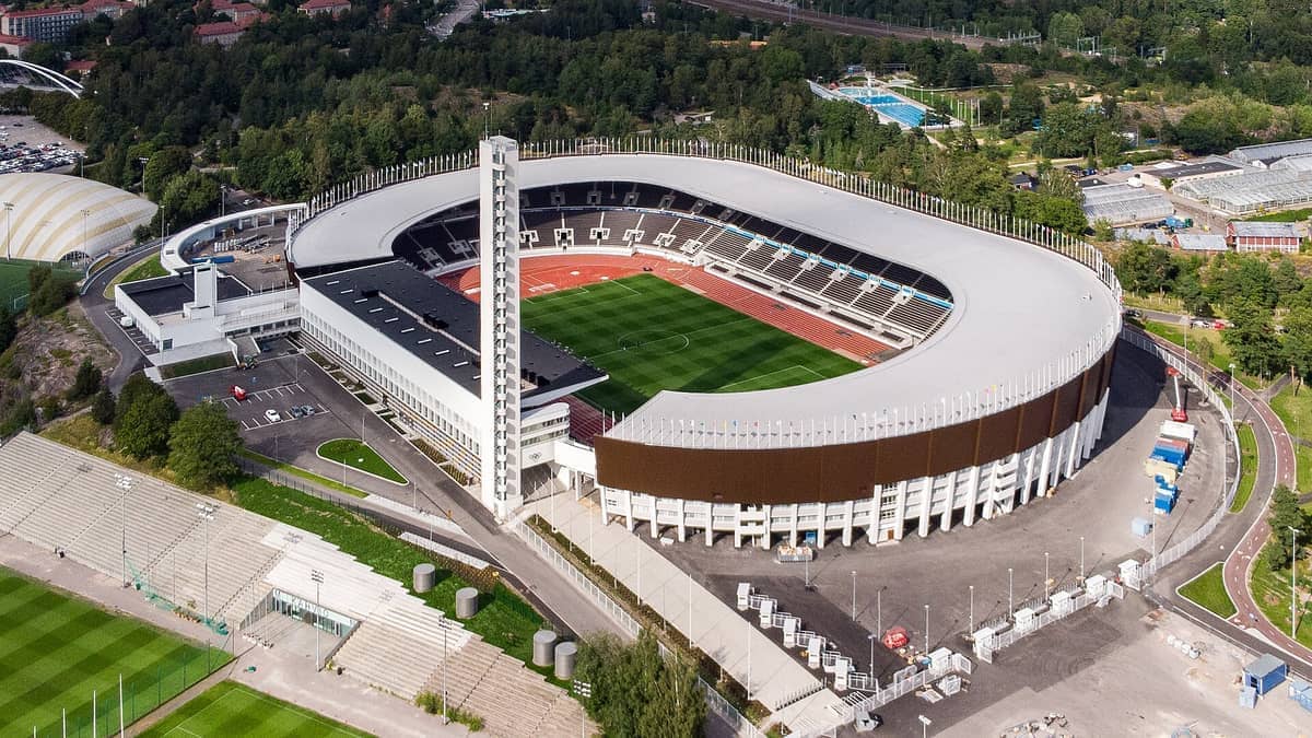 Vista aérea do Helsinki Olympic Stadium com sua icônica torre branca, campo verde, pista de atletismo e árvores ao redor em um dia ensolarado.