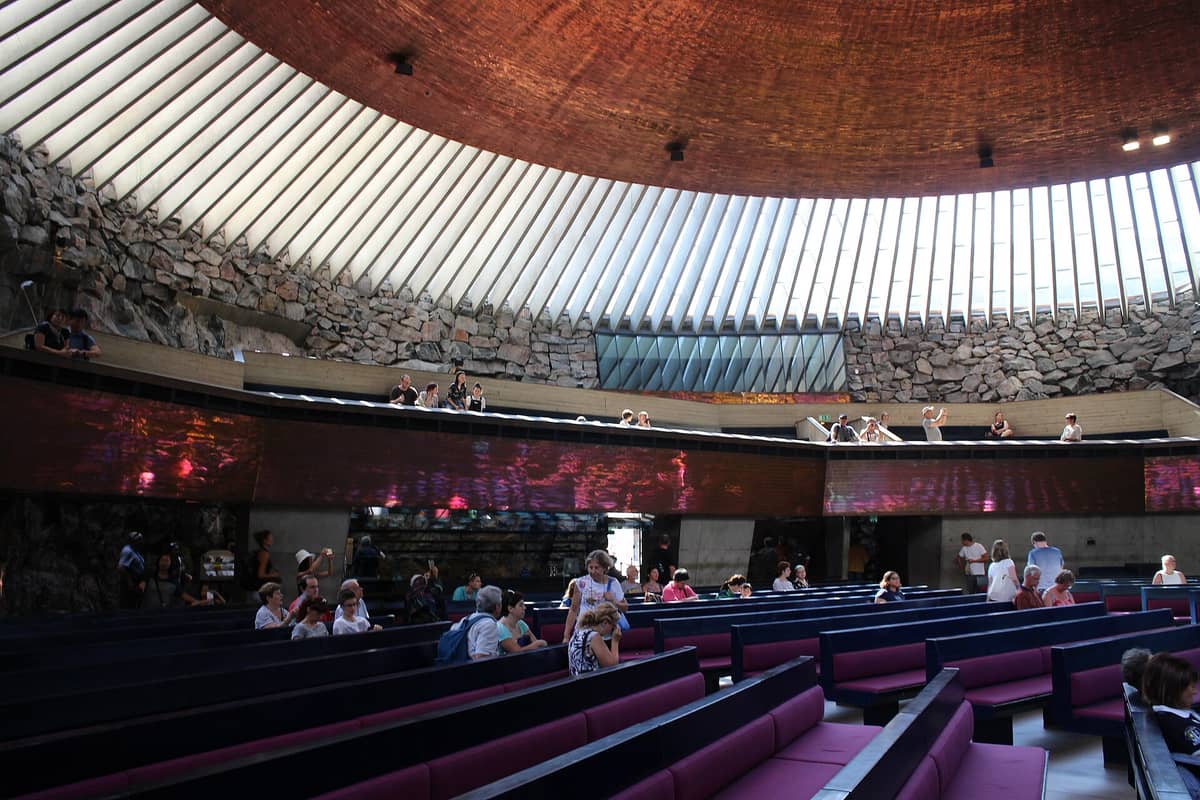 Vista interior ampla da Igreja da Rocha Temppeliaukio em Helsinki com paredes de rocha natural, altas janelas de claraboia e pessoas sentadas em bancos.