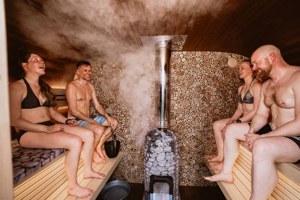 Quatro pessoas de traje de banho relaxam dentro de uma sauna revestida de madeira, com vapor subindo das pedras quentes em um ambiente moderno ao estilo finlandês.