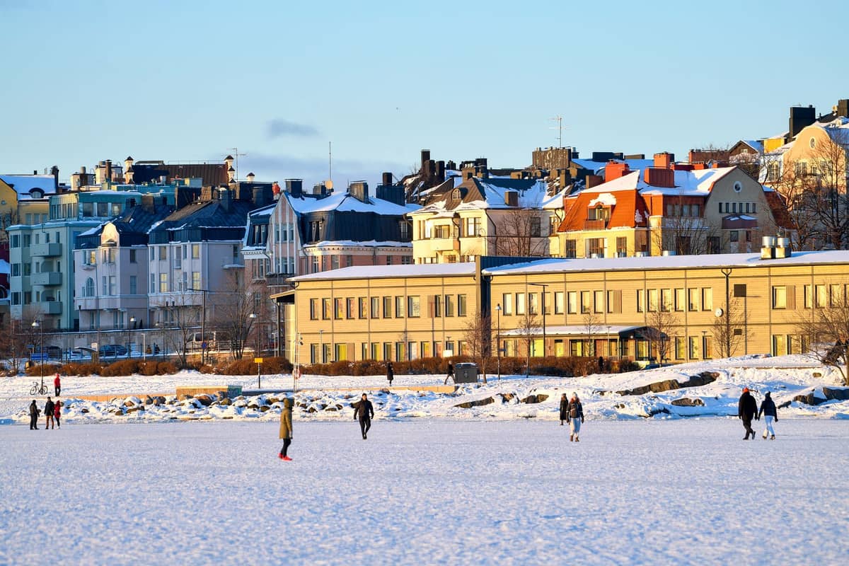 Pessoas caminhando e brincando em uma orla coberta de neve com os edifícios de Helsinki ao fundo em um dia de inverno ensolarado.
