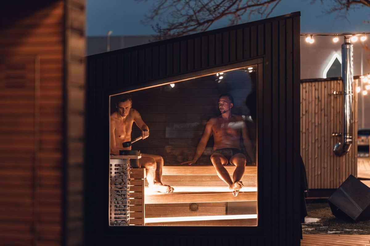 Duas pessoas sentadas dentro de uma sauna moderna de madeira, visíveis através de uma grande janela ao anoitecer em um ambiente ao ar livre em Helsinki.