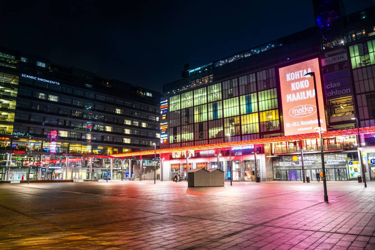 Vista noturna do complexo comercial Kamppi em Helsinki, com letreiros iluminados e uma praça quase vazia à frente.