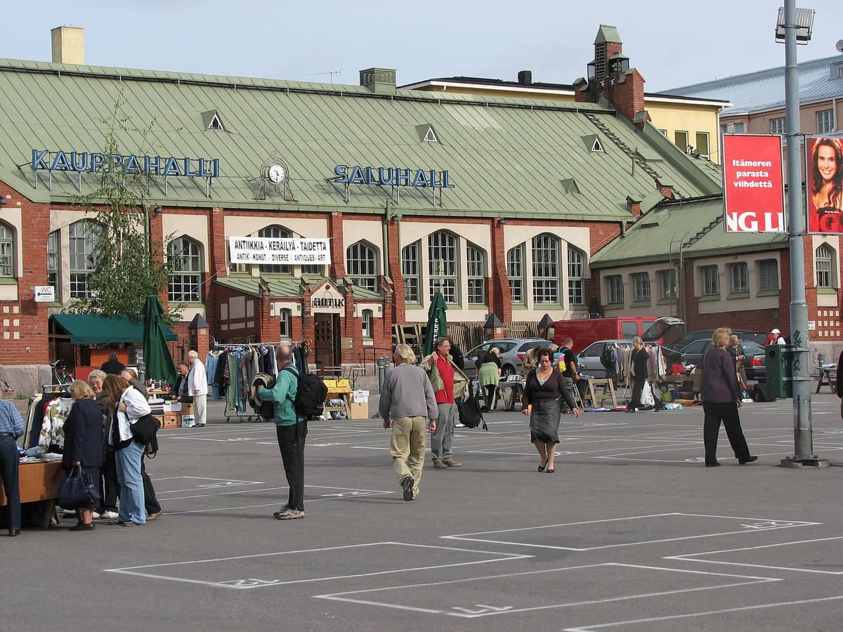 Feira de pulgas ao ar livre em Hietalahdentori em Helsinki com pessoas a examinar mesas de artigos em segunda mão em frente a um mercado coberto.