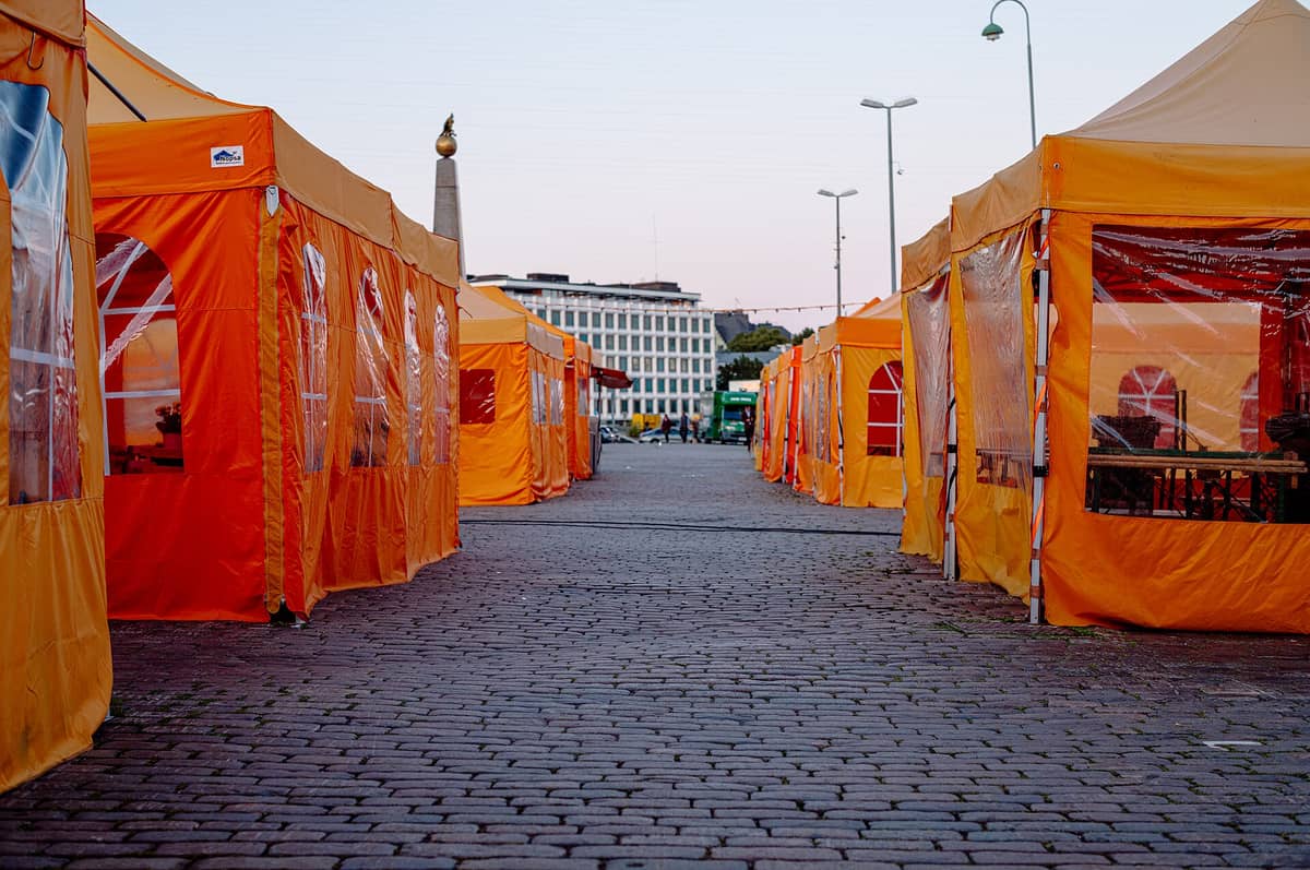 Fileiras de tendas de mercado laranja-vivas alinham uma praça de paralelepípedos em Helsinki, com um marco da cidade e edifícios modernos visíveis ao fundo.
