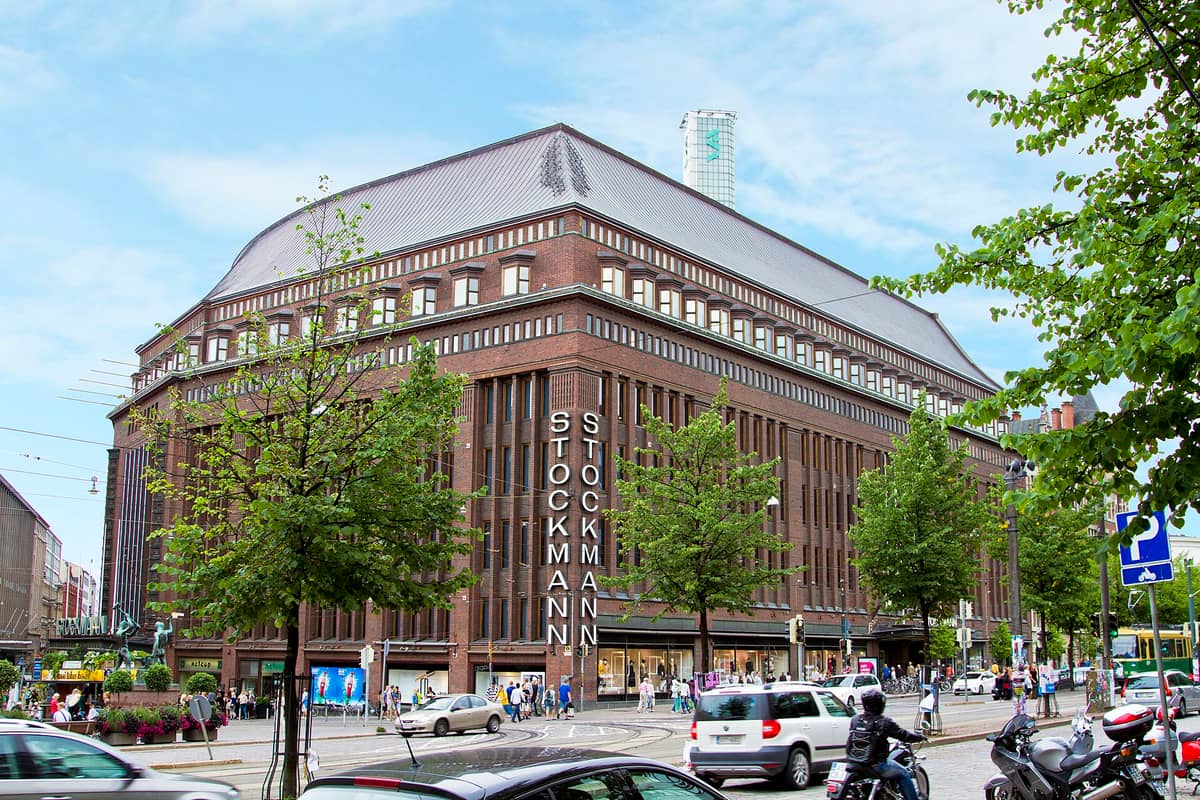 Loja de departamentos Stockmann em Helsinki, um grande edifício de tijolo com letreiros 'STOCKMANN' em destaque na esquina, rodeado de árvores verdes e tráfego intenso.