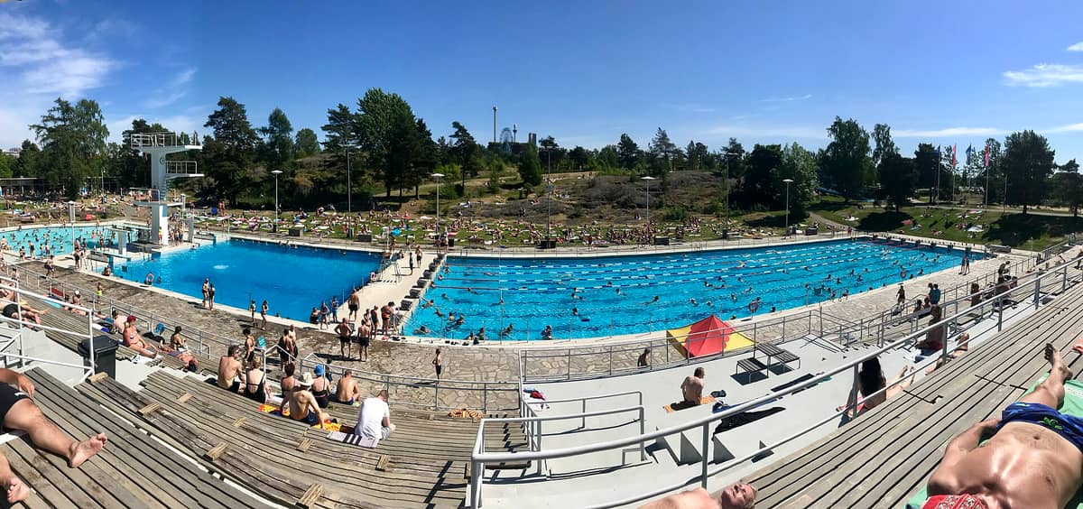 Vista panorâmica do Estádio de Natação de Helsinque mostrando as piscinas olímpicas ao ar livre, a torre de saltos, banhistas nas arquibancadas e uma grande multidão de verão sob um céu azul.