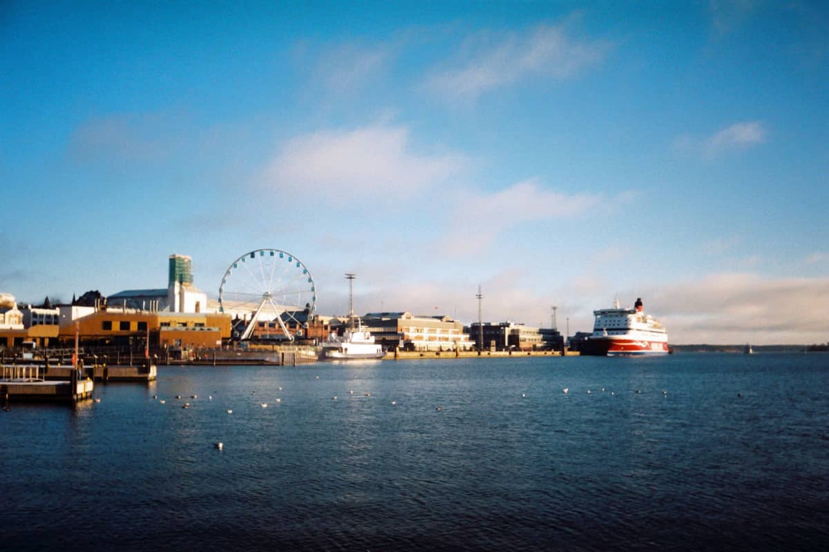 Vista do porto de Helsinki com balsas, uma roda-gigante e edifícios à beira-mar sob um céu azul.