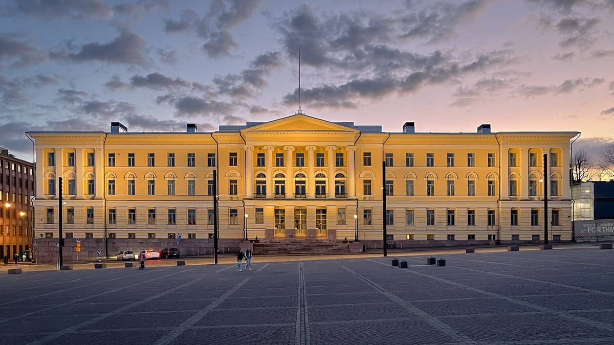 O Edifício Principal da Universidade de Helsínquia iluminado ao entardecer, visto através da vasta extensão vazia da Praça do Senado sob um céu parcialmente nublado.