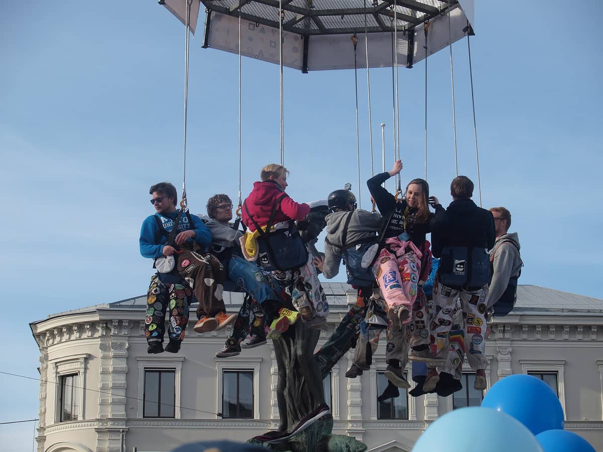 Grupo de estudantes com macacões coloridos erguidos em uma plataforma durante a cerimônia de colocação do boné em Havis Amanda em Helsinki, com céu azul ao fundo.