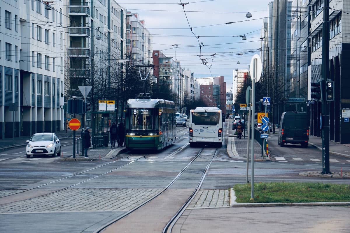 Uma cena de rua em Helsinki com um bonde verde e um ônibus em uma parada, rodeados por modernos edifícios de apartamentos.