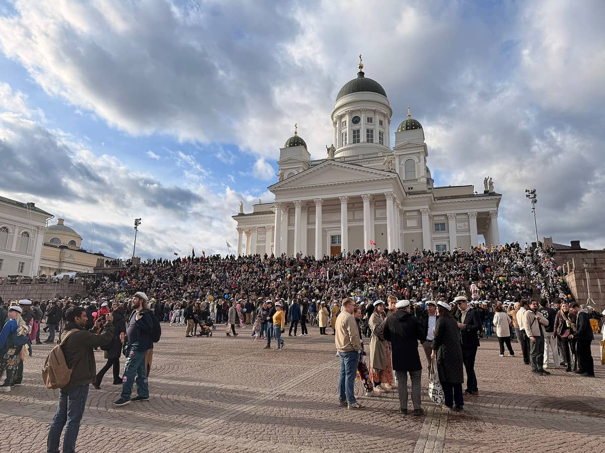 Grande multidão reunida em frente à Catedral de Helsinque para as festividades do Vappu, muitos usando bonés de estudante, sob um céu nublado.