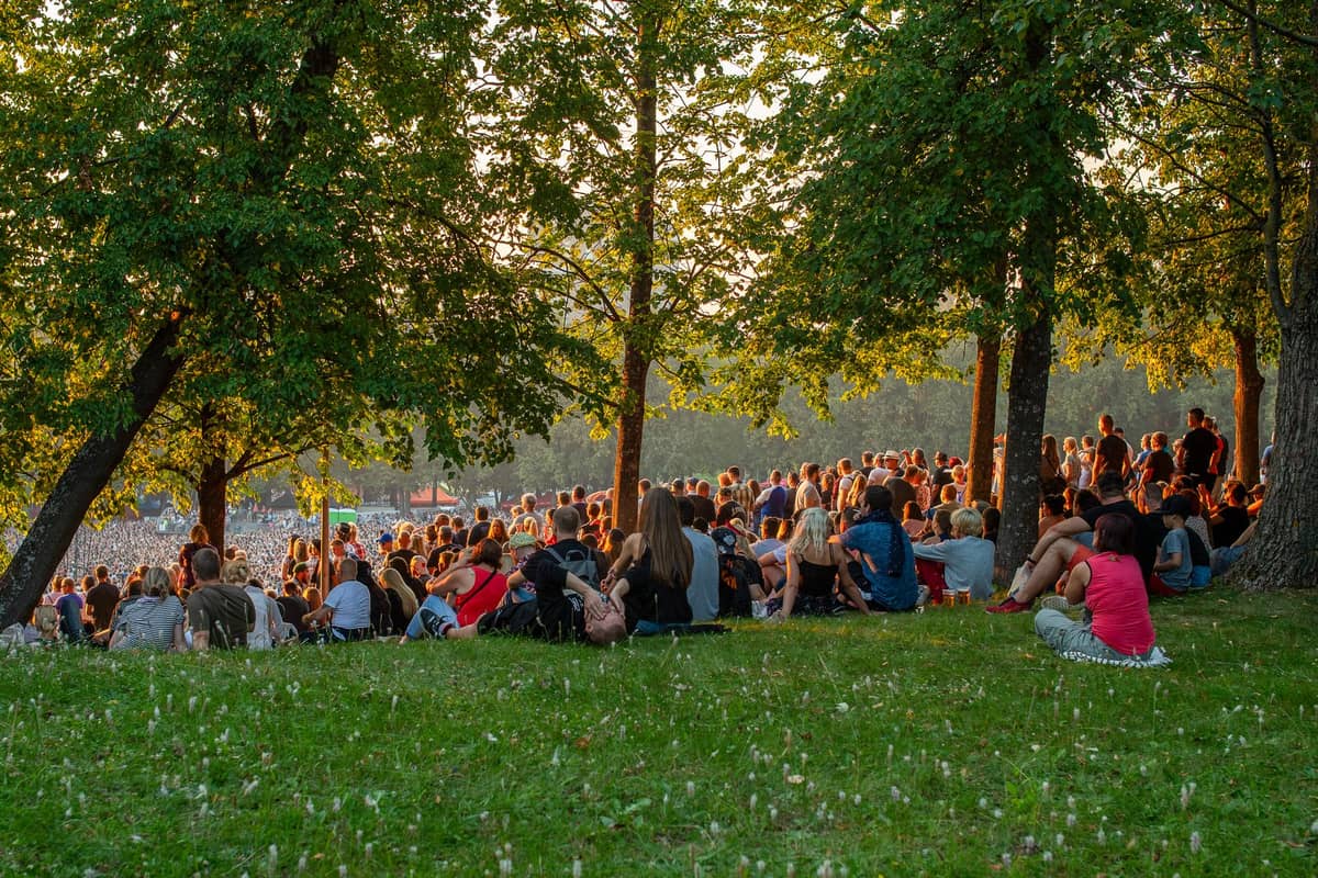 Grande multidão de pessoas sentadas na grama de um parque sob as árvores, aproveitando o sol, sugerindo um animado encontro ao ar livre ou um piquenique.