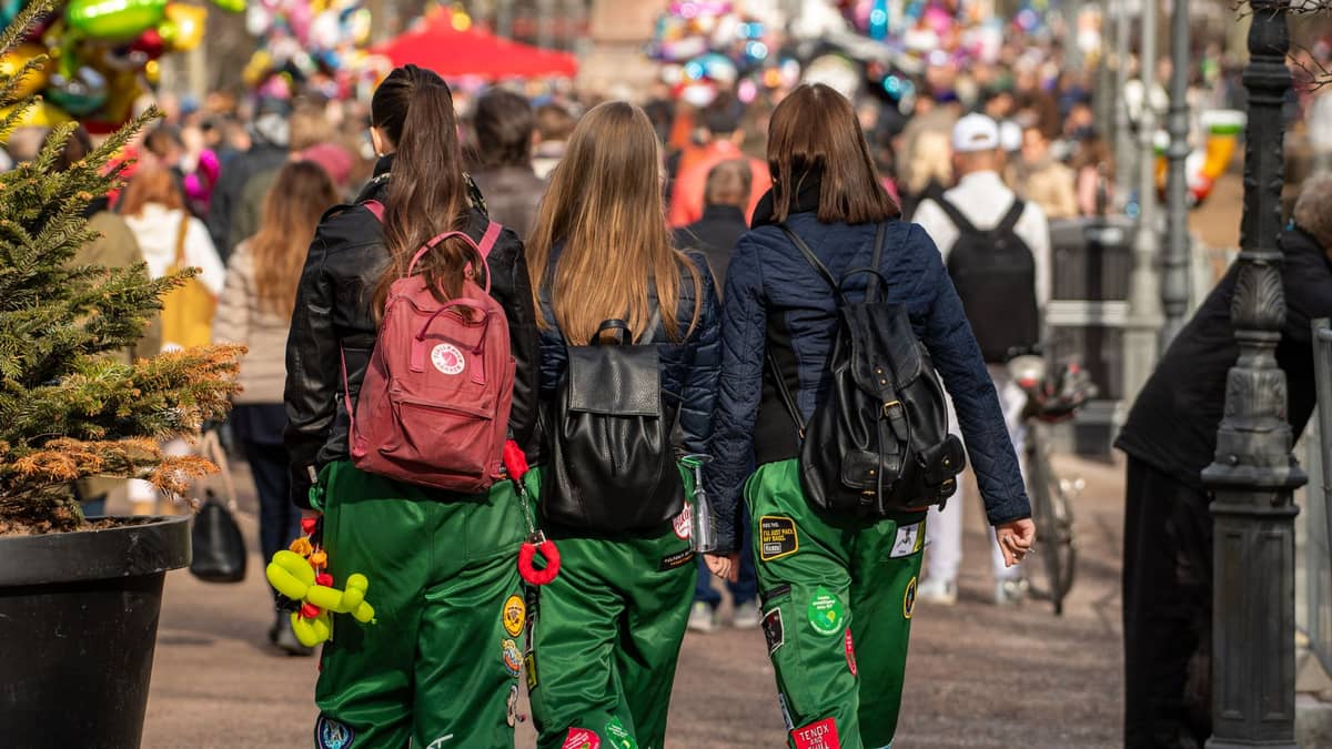 Três estudantes com macacões coloridos caminham por uma movimentada rua da cidade, rodeados de multidões e atmosfera festiva em Helsinki.