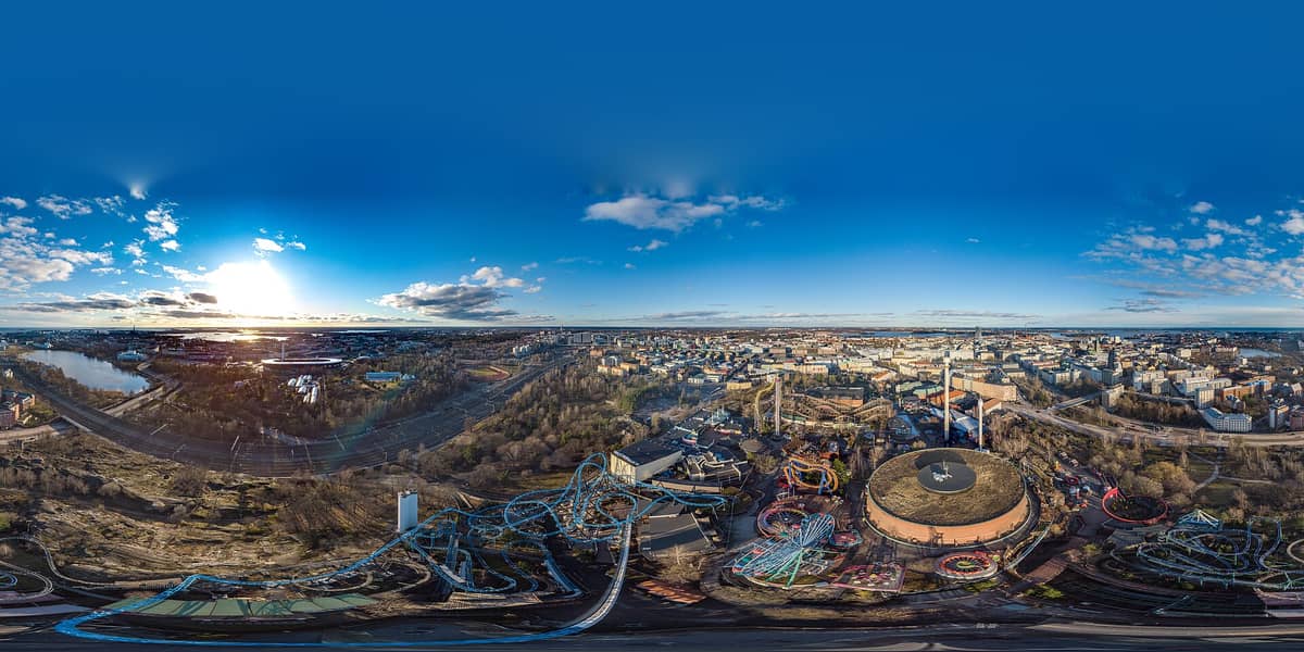 Panorama aéreo de Helsinki com o parque de diversões Linnanmäki, a icônica torre de água e a paisagem urbana se estendendo em direção ao mar sob um céu azul.