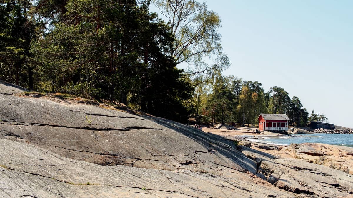 Costa rochosa e acidentada com floresta à beira-mar e uma pequena cabana de madeira vermelha junto à água em Helsinki.