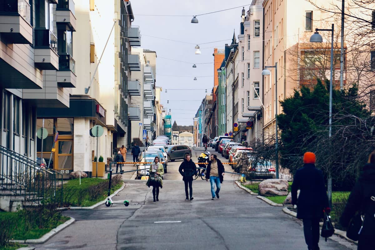 Pessoas caminhando por uma rua residencial no bairro Kallio de Helsinki, ladeada por edifícios de apartamentos e carros estacionados.